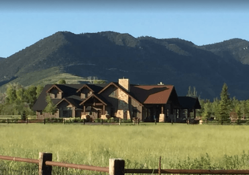 A large, rustic house with multiple roofs sits in a green field, surrounded by mountains under a clear blue sky.